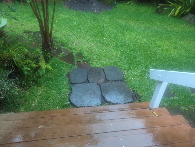 photo looking down from our front steps; old stepping stones have been removed and there are five large lava rocks placed for a new pathway, but they are wet with rain; a staked line is stretched along the side of the nascent path