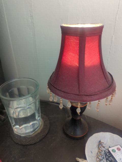 photo of a small lamp with a burgundy shade with crystal beads hanging off of it; on the same table is a glass half full of water and a small dish with knick-knacks in it