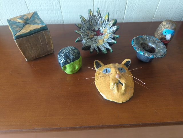 photo showing some small ceramic objet d'art: a gold box with a lid, a head with dark hair and a green face, a strangely colored flower-like thing, an orange big-cat mask with blue eyes, a small blue and gold pot/vase, and a gold-haired head with blue eye mask, a large white nose, and red lips