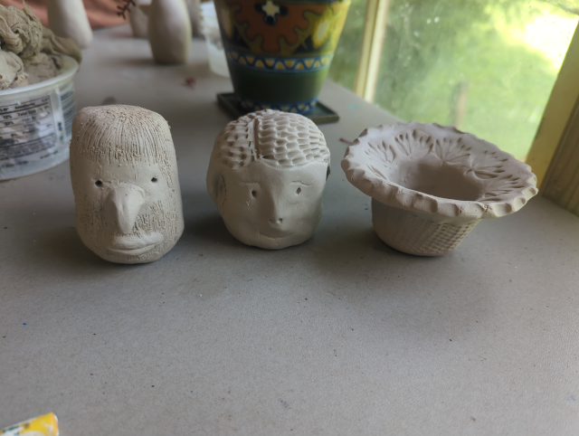 photo of three clay figures, on the left is a cylindrical face with a beaked nose, thick lips, and long, stringy hair; center is a round face with a tiny nose, sad expression, and minimal mouth; on the right is a small bowl with a scalloped edge, a flared mouth with a vegetation pattern stamped in it, and a textured base