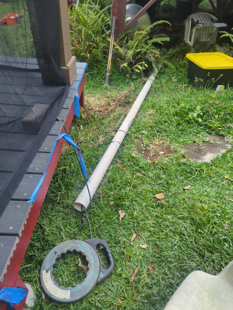 photo of conduit, before burial, with the lighting wiring being routed through it; a large-diameter white PVC pipe is visible with a fish tape and balck wire running through it, running from under the deck to under the pavilion