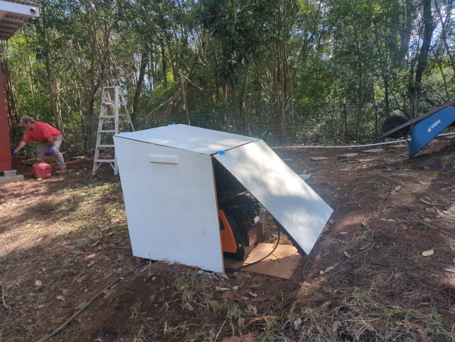photo of the generator house, now adorned with a coat of white primer; in the background, the Hired Hand is applying a coat Twoprops Compound Red to the studio