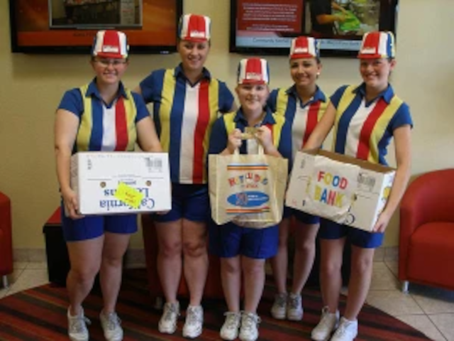 photo of a crew from Hot Dog on a Stick, wearing their signature polyester uniforms with broad red, white, yellow, and blue stripes with caps and white sneakers, carrying food bank donations