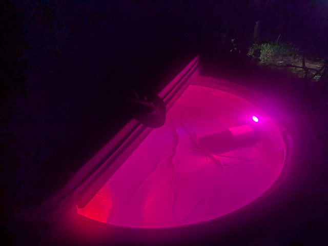 photo of the hot tub at night, with red and blue underwater lights; the cover is half on and a broad-brimmed hat rests on it