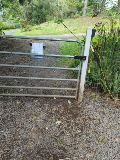 photo of our front gate from the back, showing a paper in a Ziploc bag clipped to the gate and an orange extension cord running through the fence to the driveway