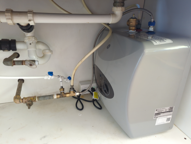 photo under the sink showing white PVC waste piping, disconnected steel hot water supply, and a cold water supply line with two tees, one line connected to plastic tubing, one connected to the faucet cold supply, and the third connected to a point-of-use hot water heater. The hot water heater is, in turn, connected to the faucet hot water supply