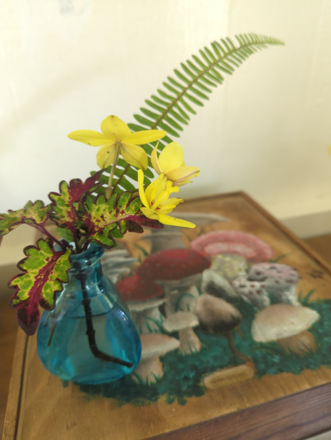 photo of a small flower arrangement with a fern frond behind yellow blossoms and a base of variegated leaves; the arrangement is placed atop a wooden box decorated with a mushroom motif