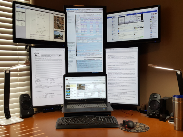 photograph of a laptop computer surrounded by many connected monitors displaying different types of documents