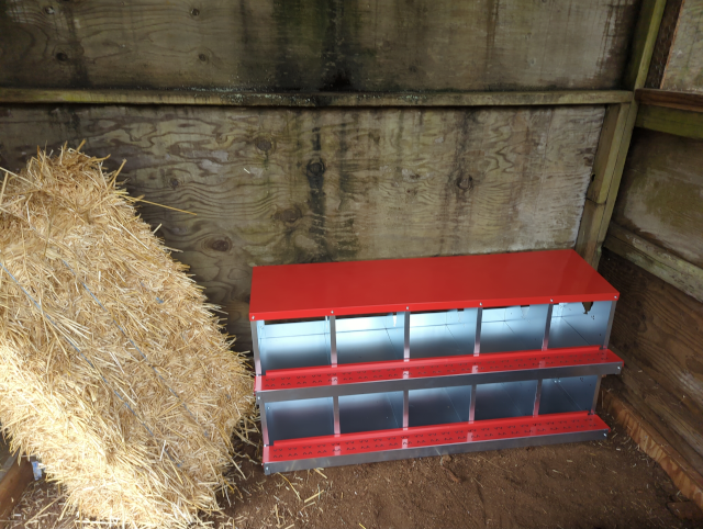 photo of a bank of ten nesting boxes made of galvanized steel with a bright red enamel roof, sitting on the ground in the henhouse