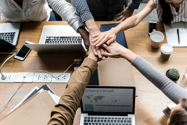 stock photo of a tabletop with some networked laptops, coffee cups, and five diverse-appearing hands reaching across the table and stacked on one another
