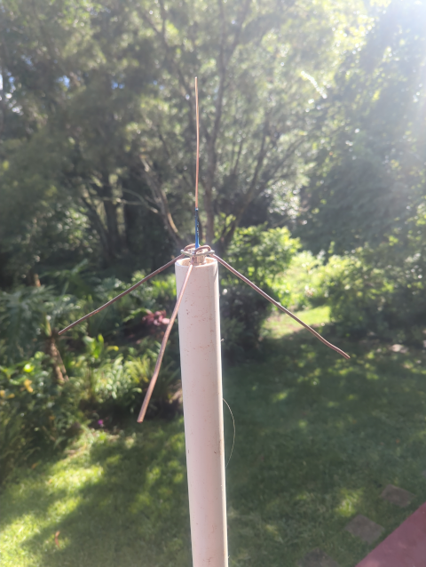 photo close-up of the quarter-wave ground-plane vertical antenna, showing the PL-259 connector upon which it is built, the vertical copper wire, and four ground-plane wires sloping off of it, all mounted on a length of PVC pipe