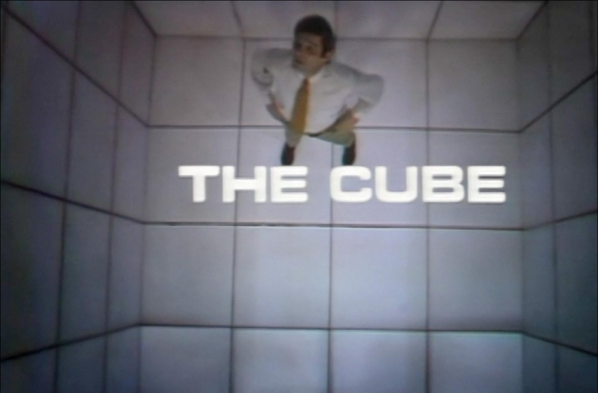 still from a television program showing a man standing in a cell made up of translucent squares, four to a side; his is wearing a tie and staring up at the camera; "THE CUBE" is in white letters across the center of the frame