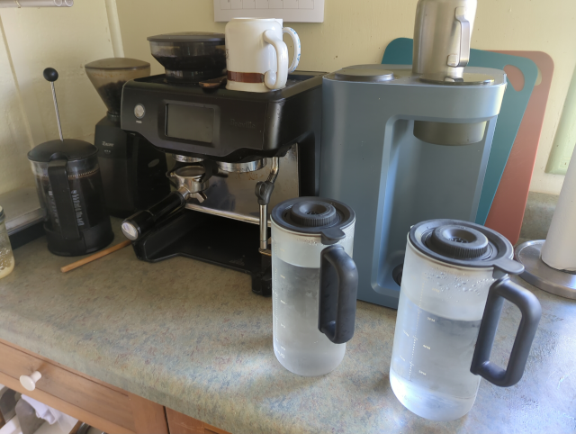 photo showing two clear glass carafes holding water in the foreground, and a tabletop water filter, an espresso machine, a press pot, and a coffee grinder in the background